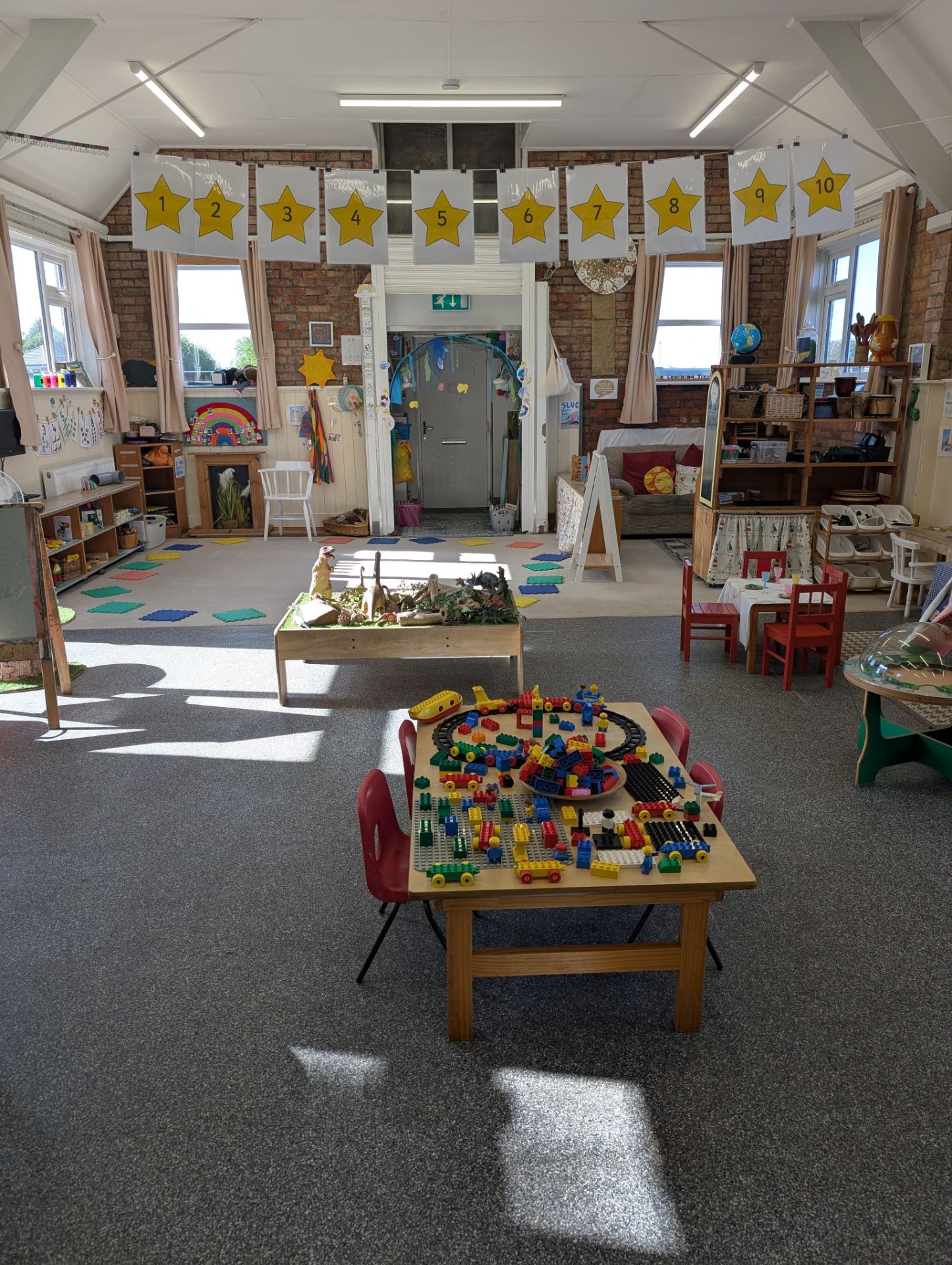 A bright, well-organized early-years classroom with large windows, exposed brick walls, and numbered star decorations hanging from the ceiling.