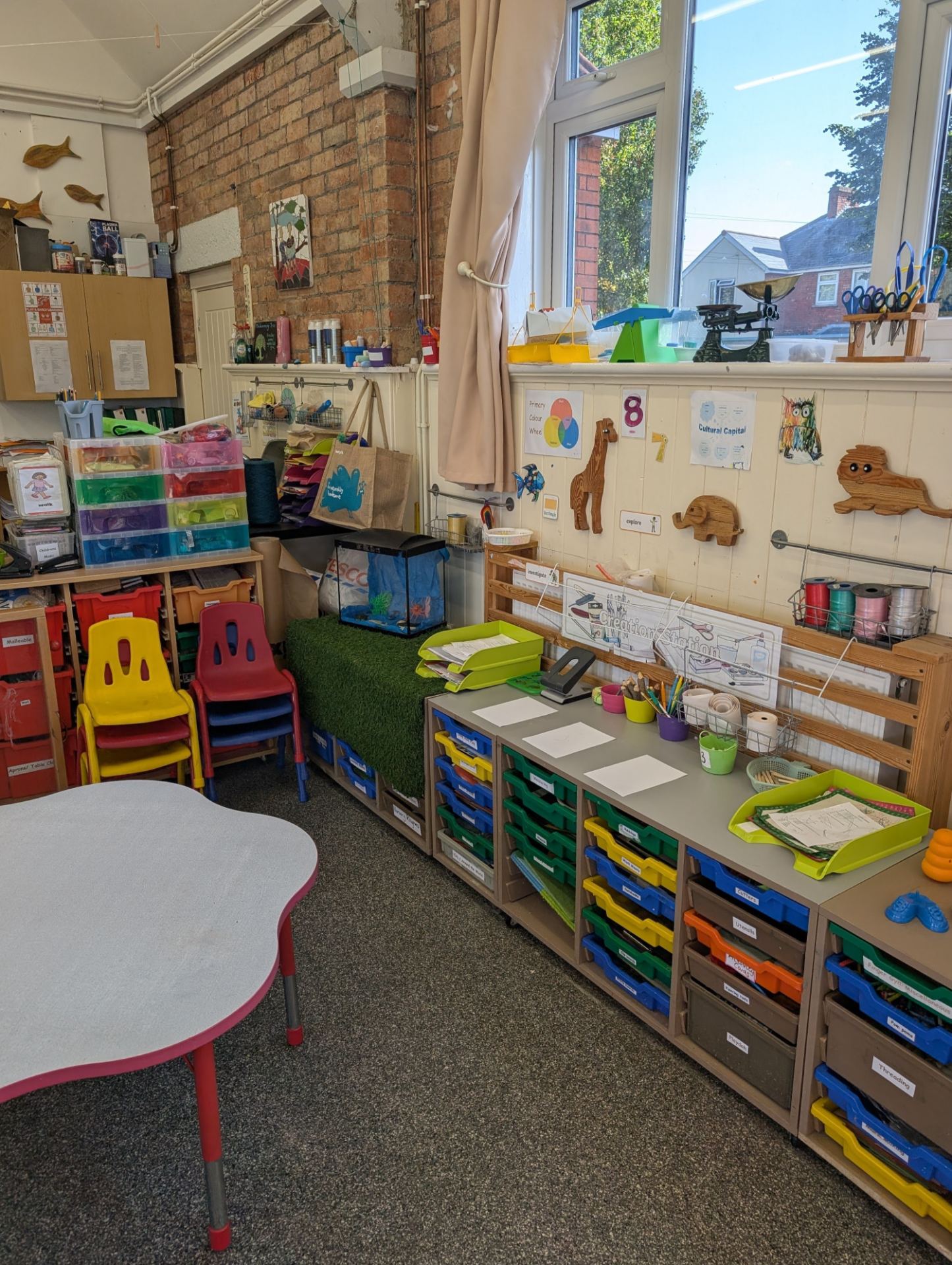 A bright, well-organised primary classroom with colourful tables, stacked chairs, storage units with labelled drawers, wall displays, and large windows overlook