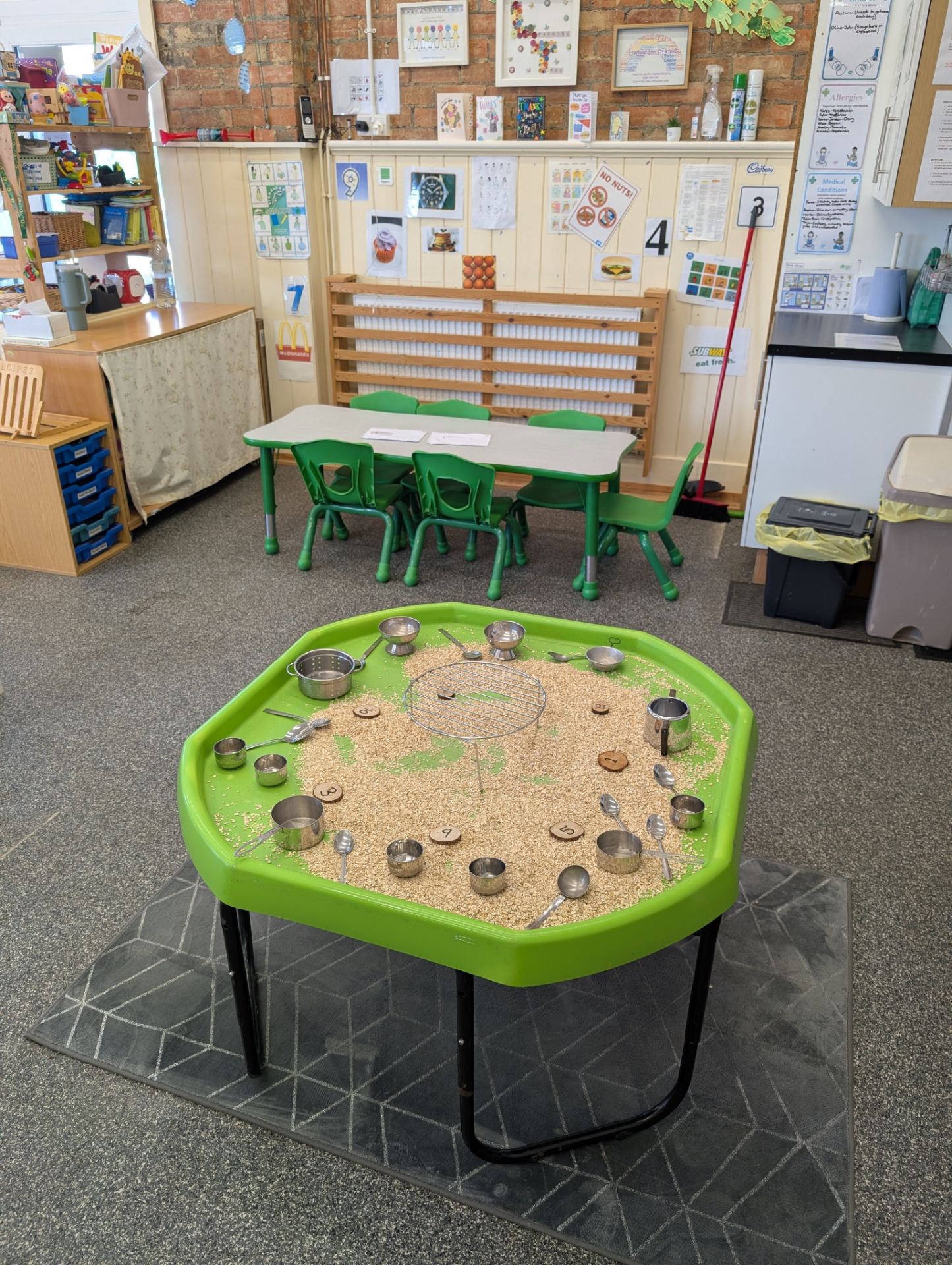 A bright, well-organized classroom with a prominent sensory sand table in the foreground featuring metal measuring cups and natural materials arranged in a circ