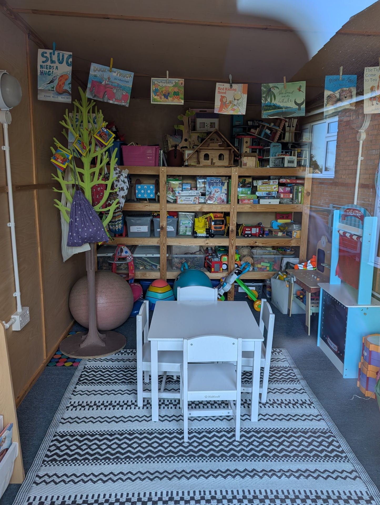 A well-organised early-years classroom or activity room with child-sized furniture, shelving stocked with toys and learning materials, colourful displays includ